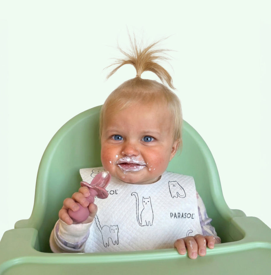 Baby in a high chair with a messy face, holding a spoon and bib.