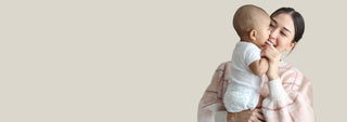 A woman in a pink plaid blanket holds and kisses a baby wearing a white outfit, set against a solid light beige background with ample negative space on the left.
