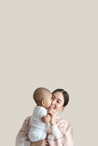 A woman holding a baby against a solid light beige background. The woman has dark hair and is wearing a light pink and white plaid cardigan, while the baby is dressed in a white t-shirt and diaper.
