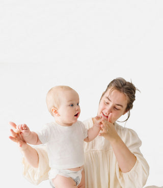 A woman in a cream-colored blouse holds and supports a baby wearing a white t-shirt and diaper, set against a solid white studio background.
