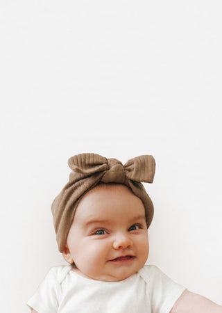 A smiling baby wearing a brown corduroy bow headband and a white short-sleeve bodysuit, photographed against a plain white background.
