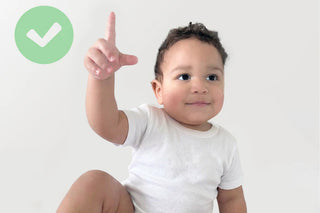 A baby wearing a white short-sleeve bodysuit sits against a light gray background, pointing upward with one finger. A green circle icon with a white check mark appears in the top left corner.
