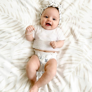 A baby lying on a white leaf-patterned blanket, wearing a white short-sleeve crop top, a white diaper with gray butterfly prints, and a black-and-white polka dot bow headband.
