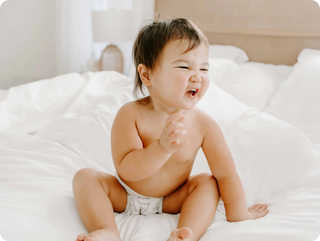 Baby sitting on a white bed wearing a diaper, smiling.
